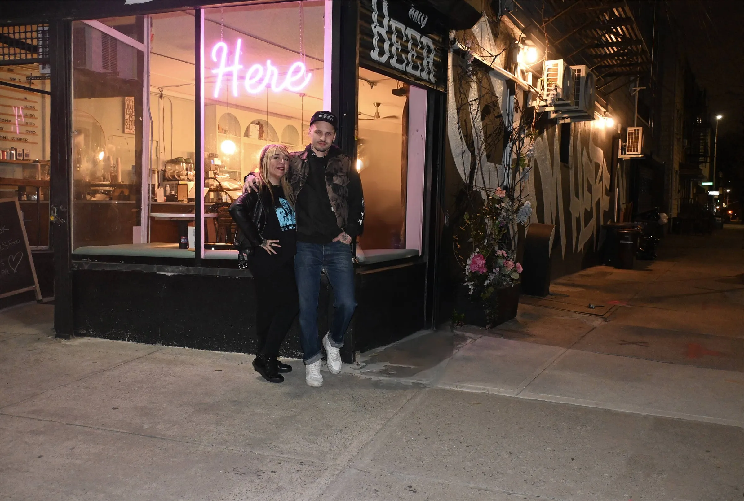 Two people stand close together on the sidewalk at night in front of a corner storefront. A pink neon sign reading Here glows in the window behind them, and a large black and white painted mural covers the wall to the right.